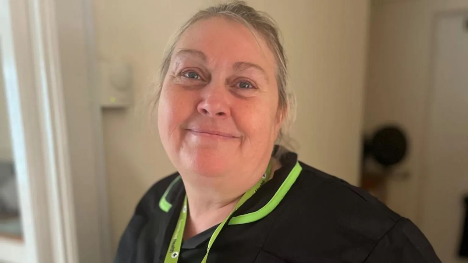 Kellie is smiling at the camera. She has blonde hair tied back. She is wearing a black work tunic that has like green trims.