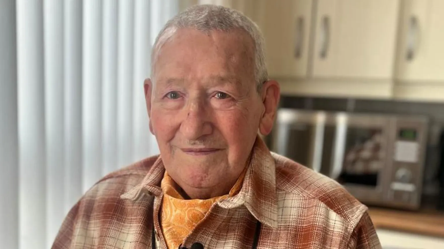 Alan is looking at the camera. He is wearing an orange checked shirt with an orange neck scarf. He has short grey hair.