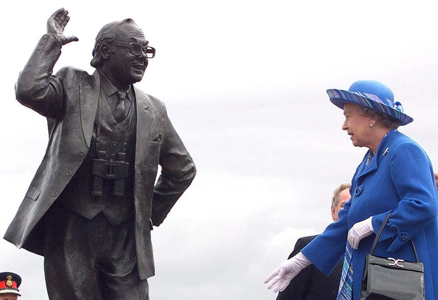 Queen Elizabeth II, dressed in blue coat and hat, is standing near a statue of Eric Morecambe