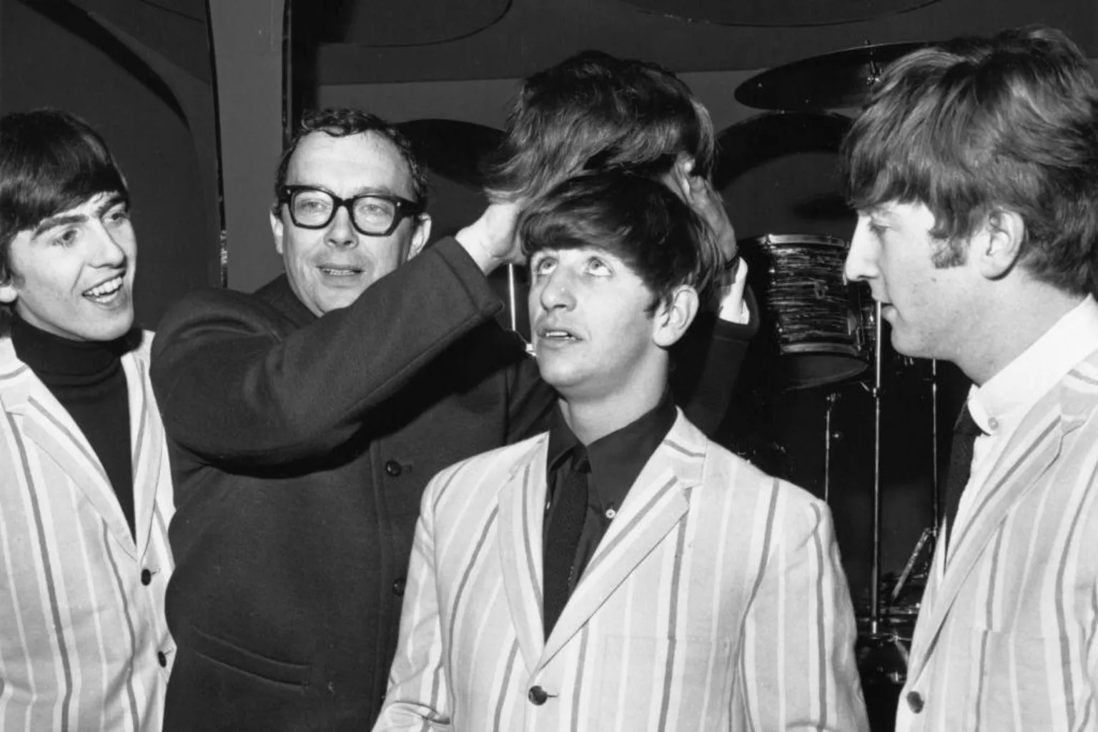 Eric Morecambe is dressed in a Beatles-style coat and holding a Beatles-style wig above the head of Ringo Starr as George Harrison and John Lennon look on from either side.