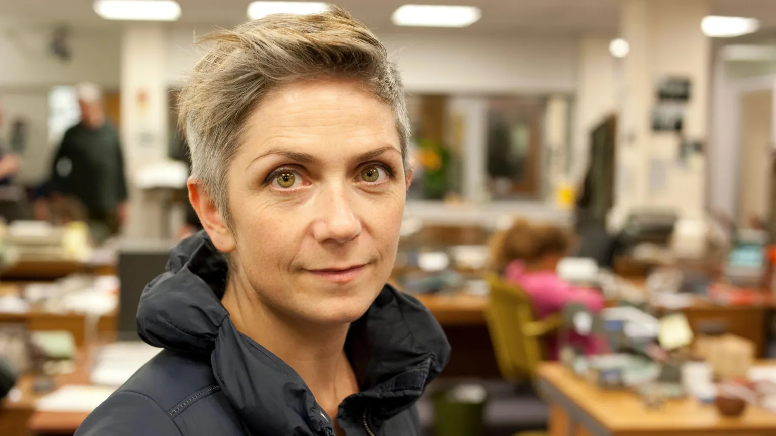 A photo of author Denise Mina from the shoulders up with the background blurred. She has short grey hair, with a hint of blonde on the ends. Denise looks straight at the camera with a small smile on her face. She wears a black puffer jacket and has hazel coloured eyes.