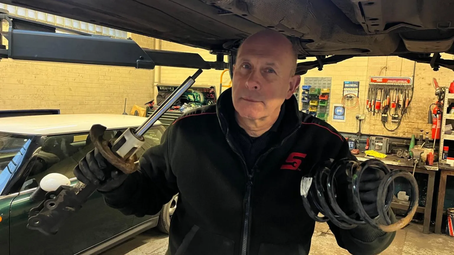 Mick Hampshire pictured under a car holding a spring in one hand and an instrument in the other.