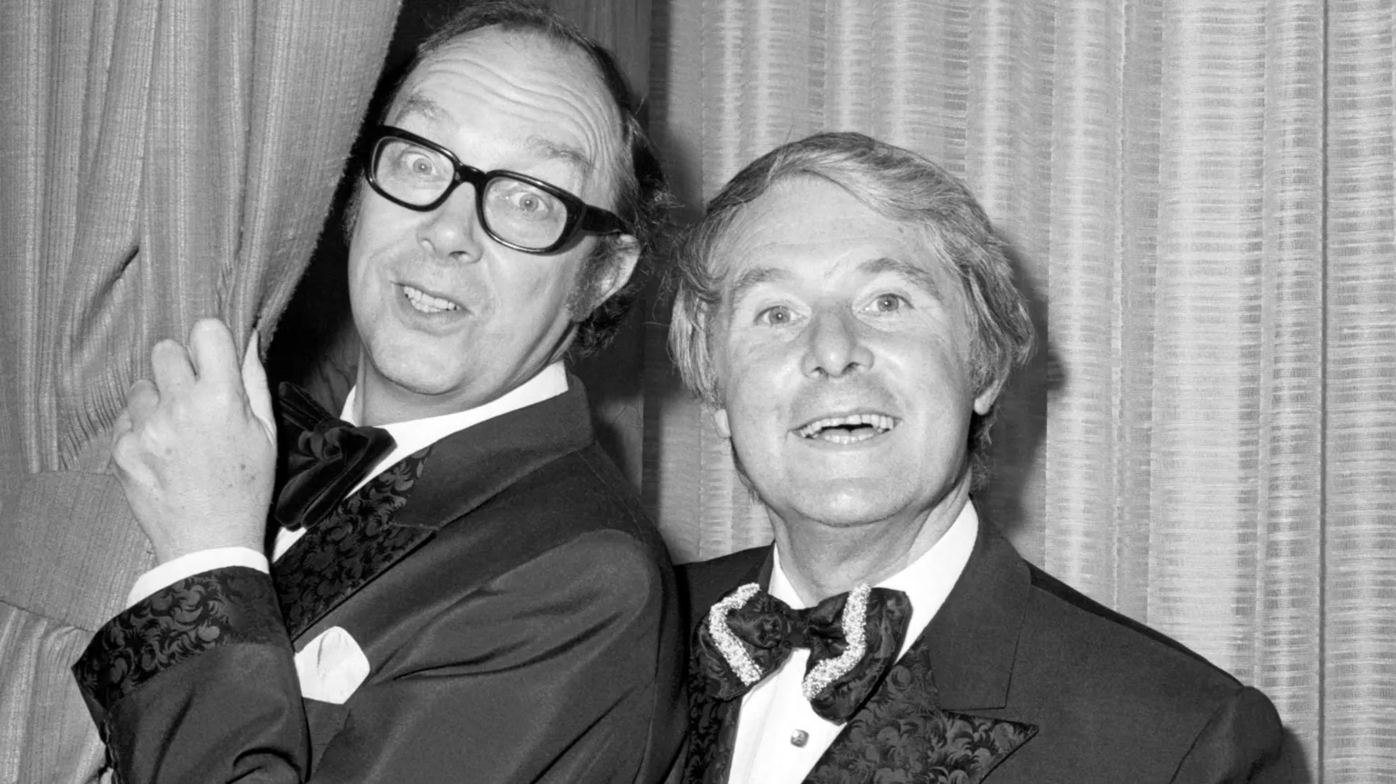 PA In a black and white photograph, the two comedians smile as Morecambe peaks out behind a curtain. They are both wearing tuxedos with large bow-ties in this backstage photograph.