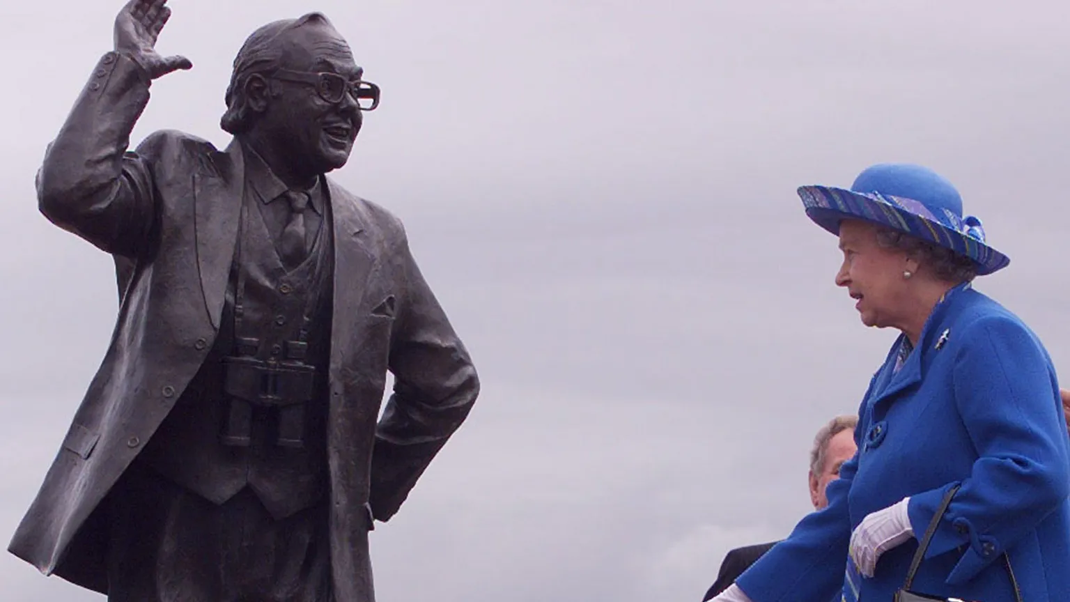 PA Elizabeth II wears a royal blue hat and coat with white gloves. She stands next to a large dark metal statue of Eric Morecambe with one hand in the air and smiling.