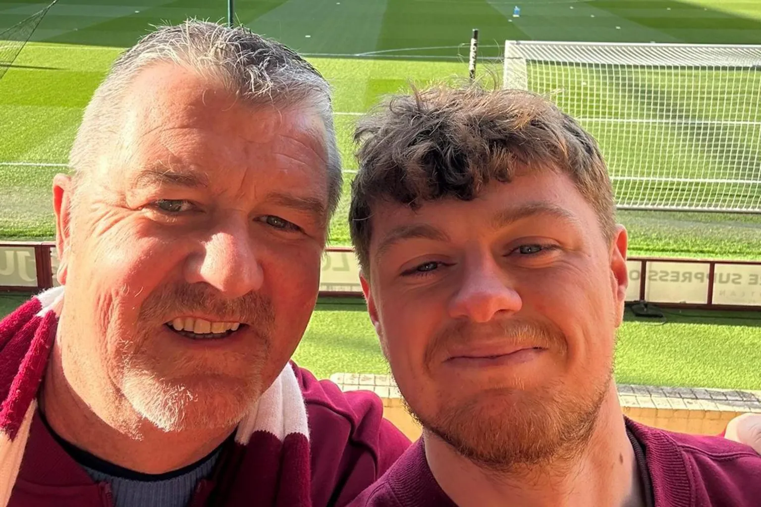 Mark Speakman Mark Speakman and his son Kyle at Tynecastle. They are both wearing maroon Hearts shirts and smiling at the camera. An empty football pitch with a corner of the goal is in the background.