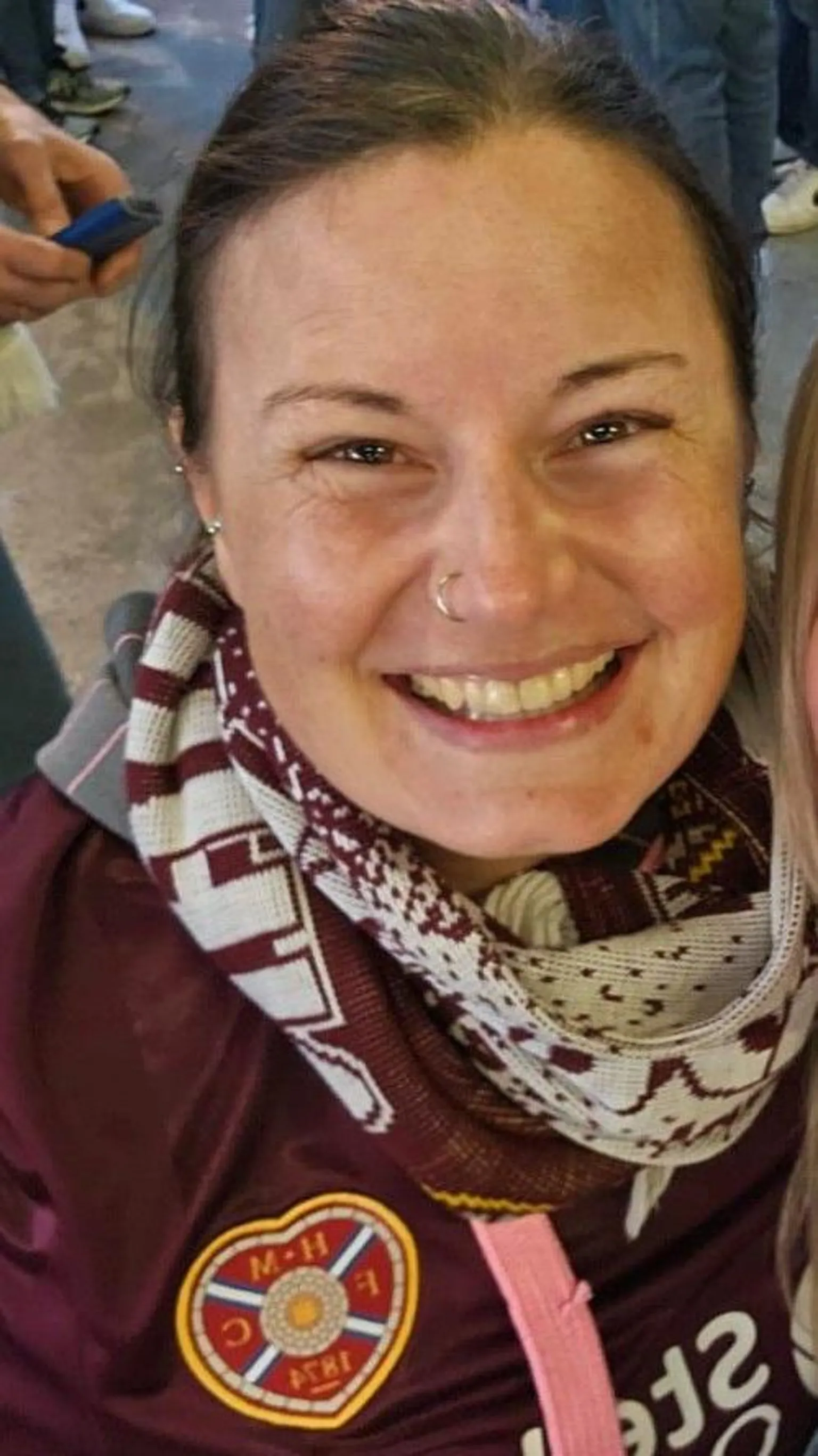 Jenny Addison Jenny Addison wearing a maroon Hearts shirt and scarf smiling at the camera.