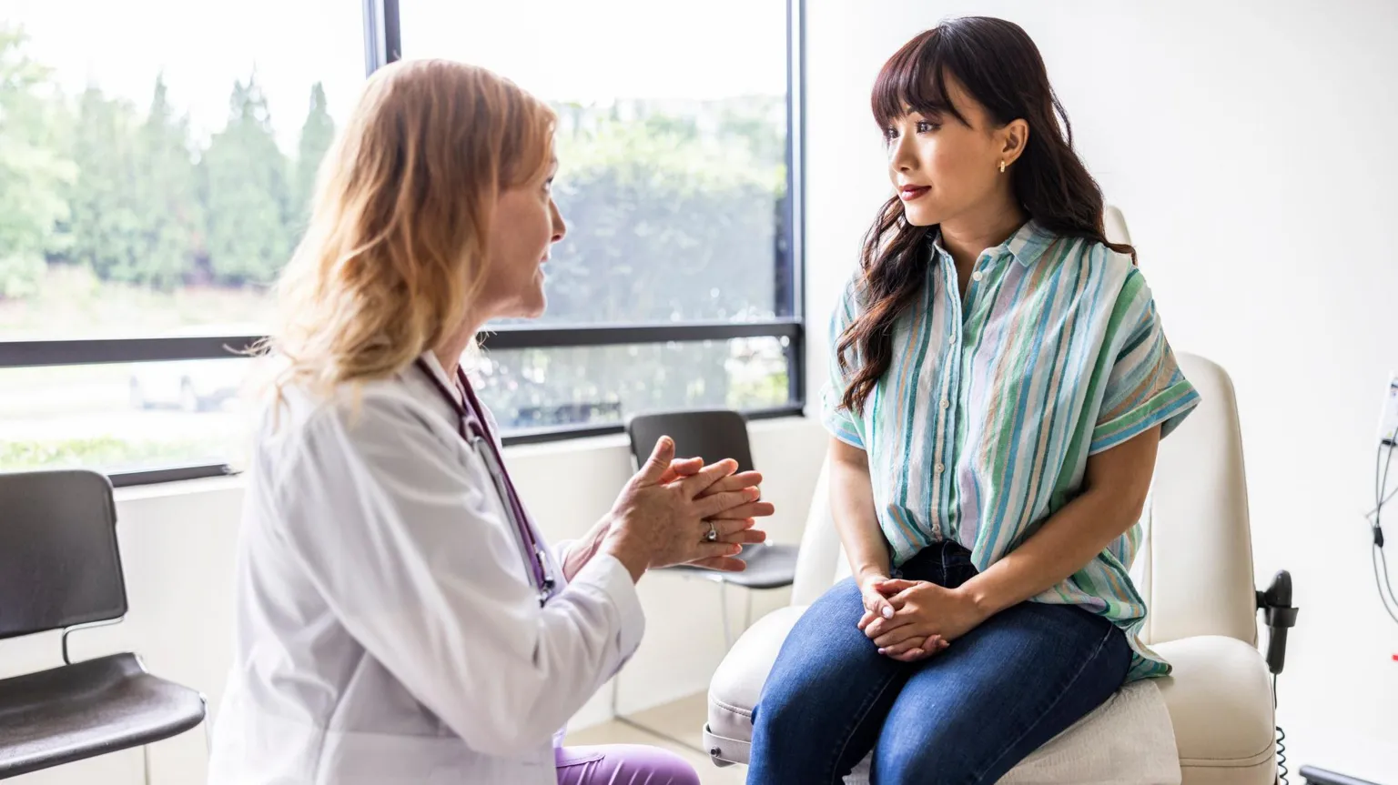  Female doctor talking with young woman in exam room. The doctor is wearing a white lab coat and pink scrubs. The woman is wearing a blue shirt and jeans and sitting on a raised exam chair. Outside the window there are trees. 