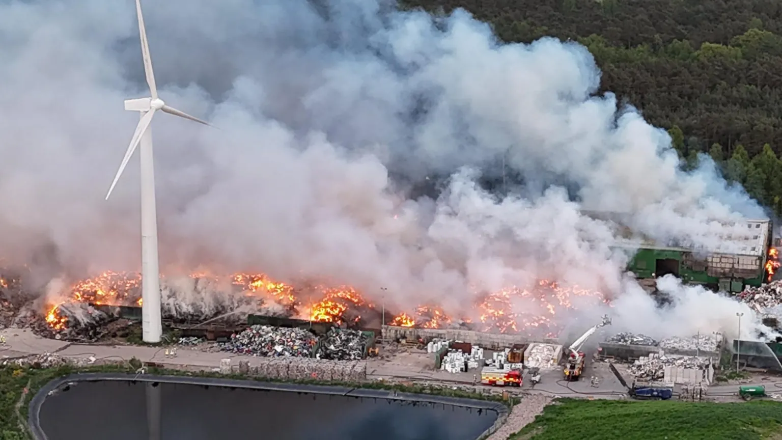  Flames spread across a large area of landfill that is next to a wind turbine. The fire is creating lots of smoke