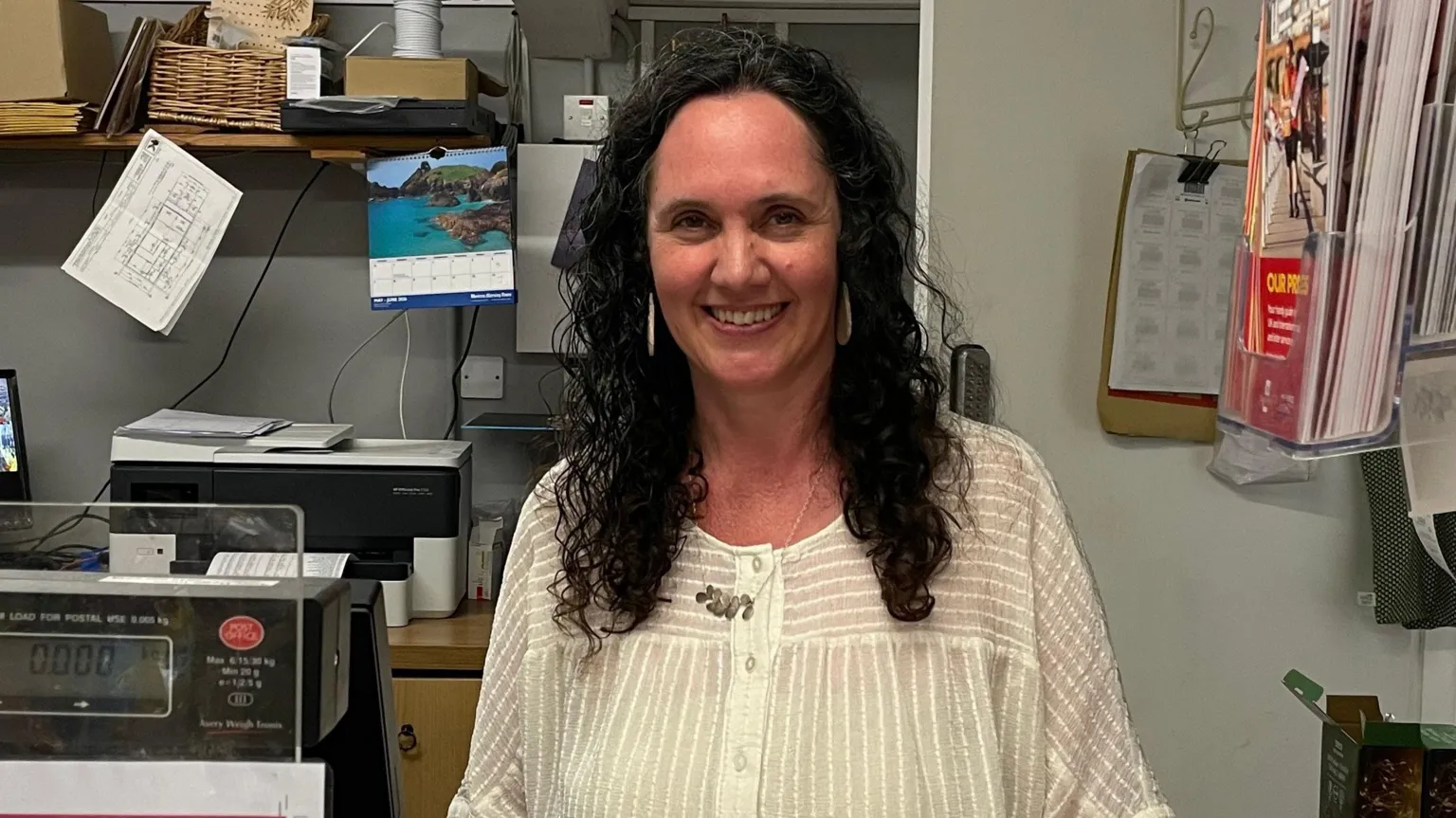 Postmistress Joanna Bickersteth stood at the Post Office counter. She has long dark hair and is wearing a white top. She is smiling straight at the camera. There is an office in the background with paper work and a calendar hung up. 