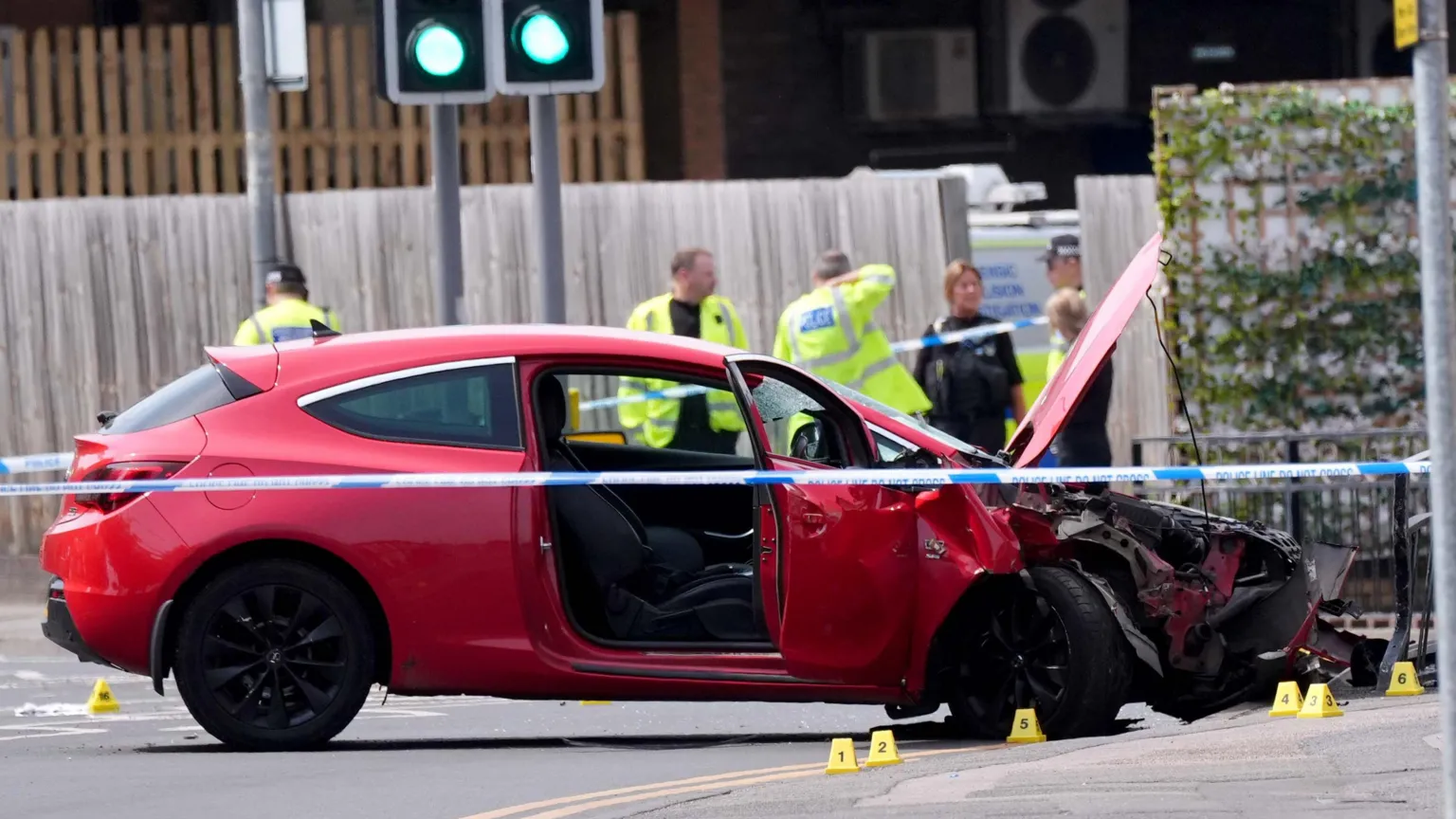  A red car that has crashed has police tape around it. Police officers in the background are wearing neon yellow hi-viz jackets 