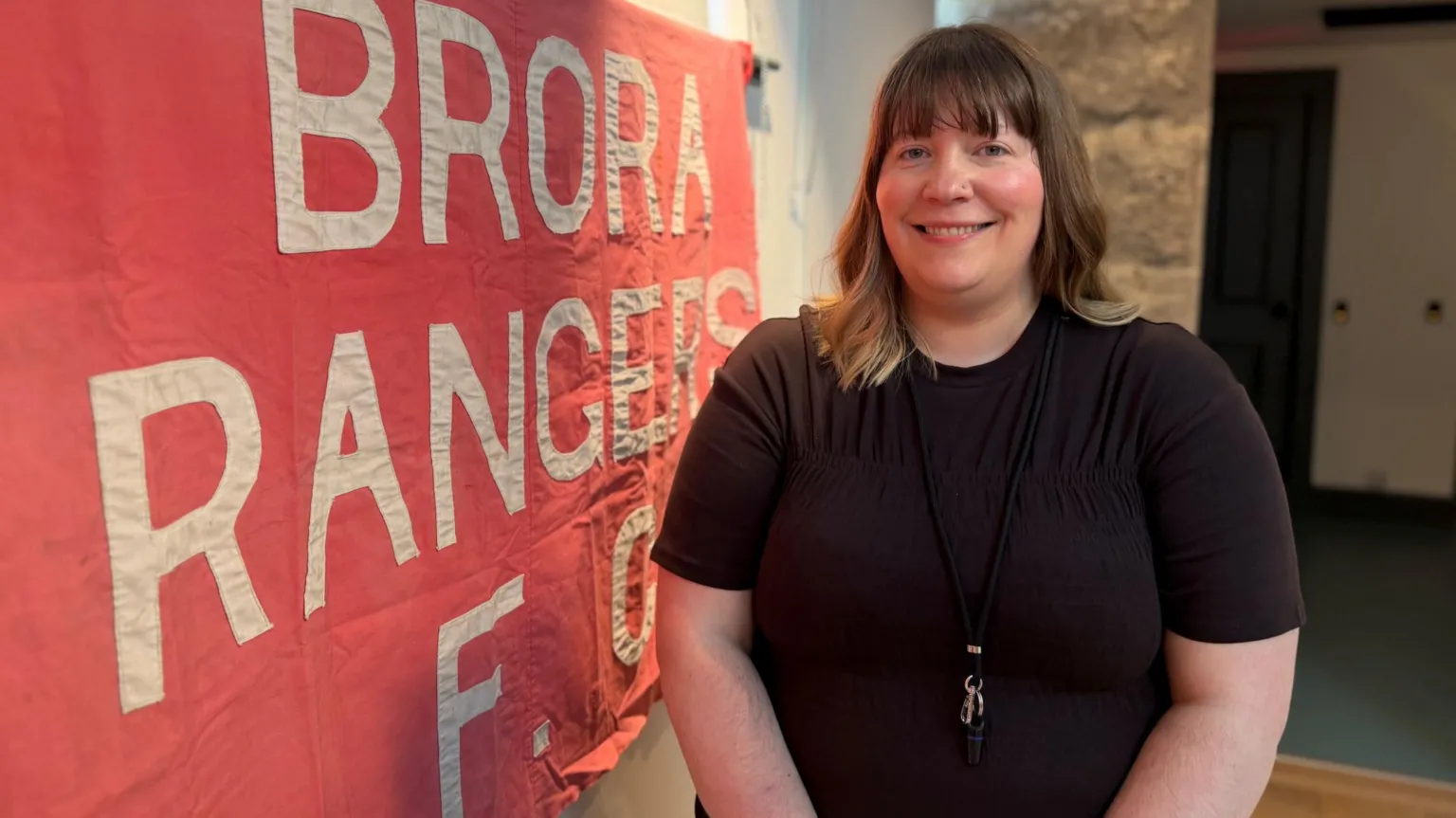 Aileen Ogilvie is standing inside Brora Heritage Centre next to a red cloth banner with the words