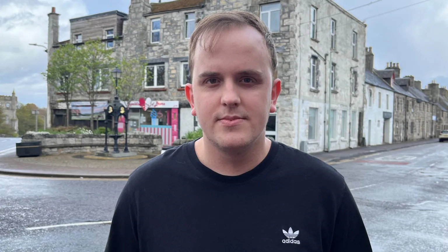 George Mackenzie is standing outside. There is a large stone building behind him. George has fair hair and is wearing a black top.
