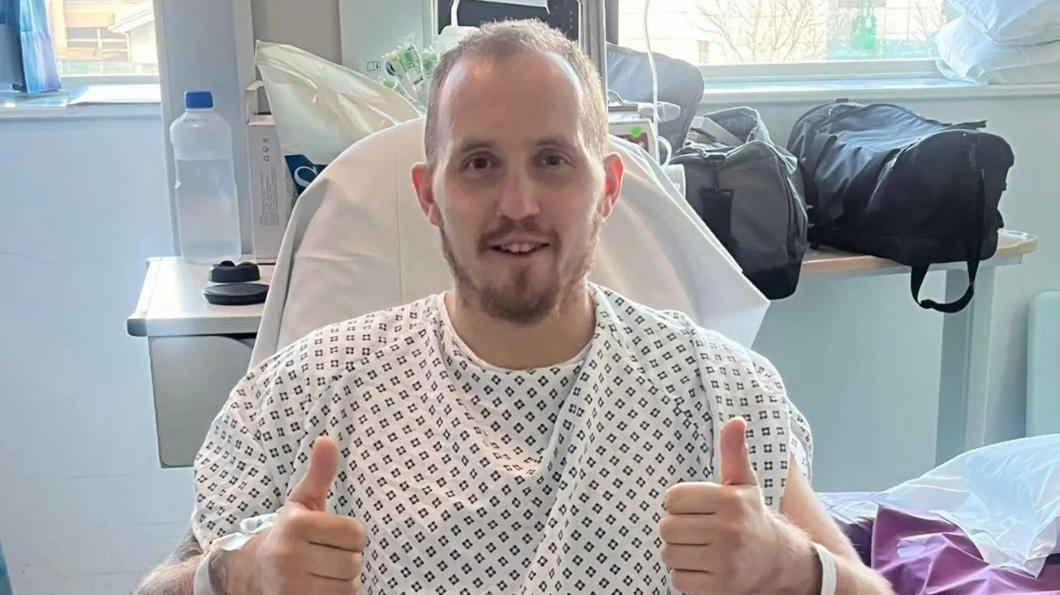 Keenan Acton Keenan is sitting in a chair in hospital smiling with both his thumbs up. He is wearing a white hospital gown with a small navy squares pattern. He has short brown hair and a beard. Behind him is a table with various bags piled on it and a window.