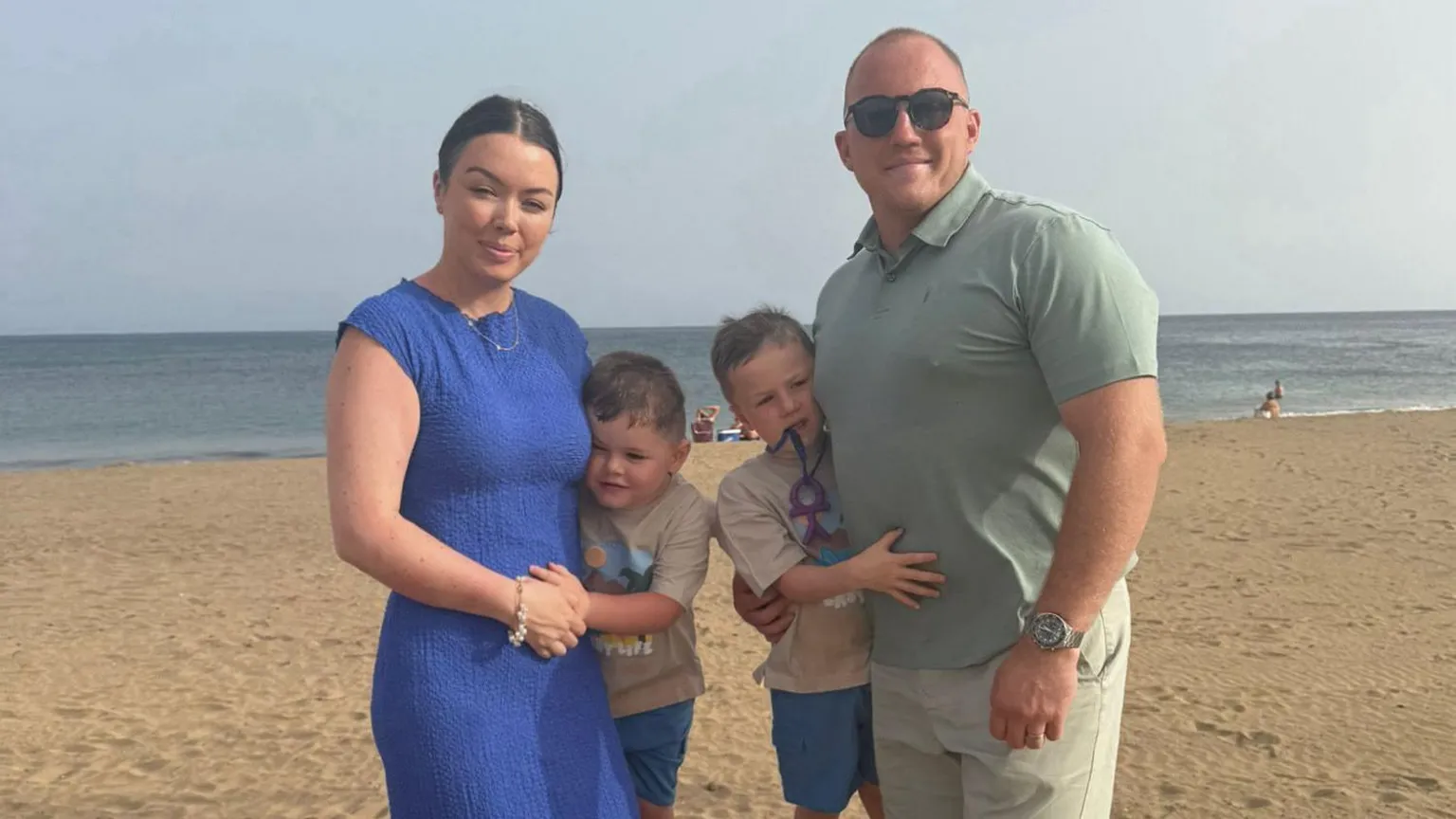 Keenan Acton Keenan, Olivia - who has tied-back brown hair - and their two young sons are wearing summer clothes and standing on a sandy beach with the sea behind them. All are smiling and looking at the camera. Olivia's dress is blue, Keenan is wearing a green polo shirt, beige shorts and sunglasses, and stood between them their boys are wearing matching beige t shirts and blue shorts.