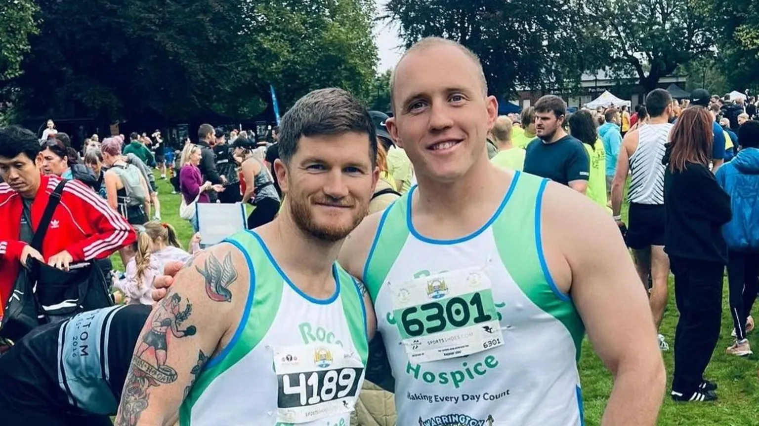 Keenan Acton Keenan (right, with very short fair hair) and a friend (who has longer, dark hair and facial hair, and tattoos all down one arm) at a 10k run event. Both are wearing matching green, blue and white running vests with race numbers pinned on them, and are smiling at the camera. Behind them are lots of other people at the event.