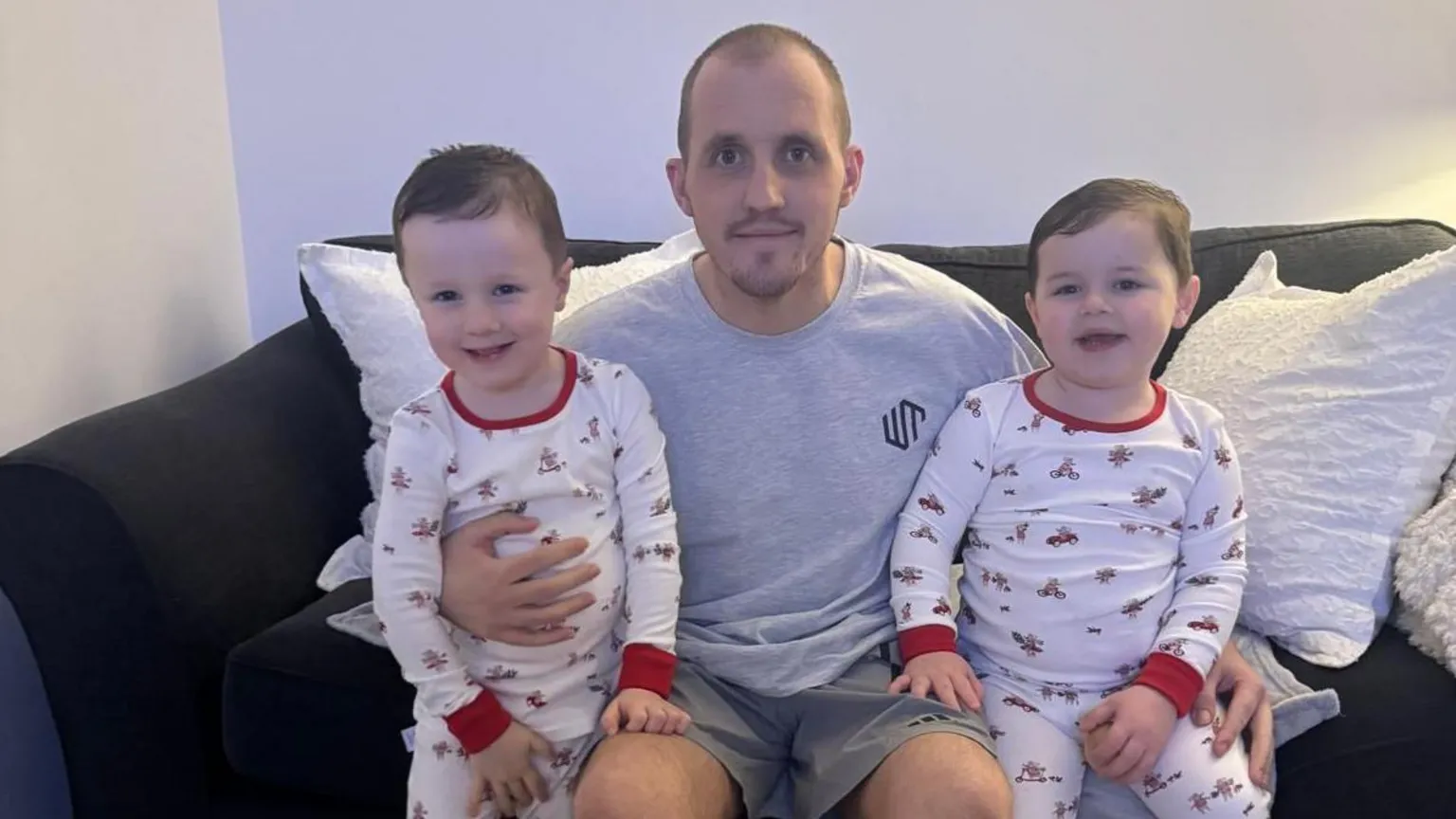 Keenan Acton Keenan is sitting on a dark sofa with his young sons sitting either side of him. His sons are wearing matching white pyjamas with red cuffs with a pattern of bikes and cars. Keenan is wearing a grey t-shirt and shorts and all are smiling.