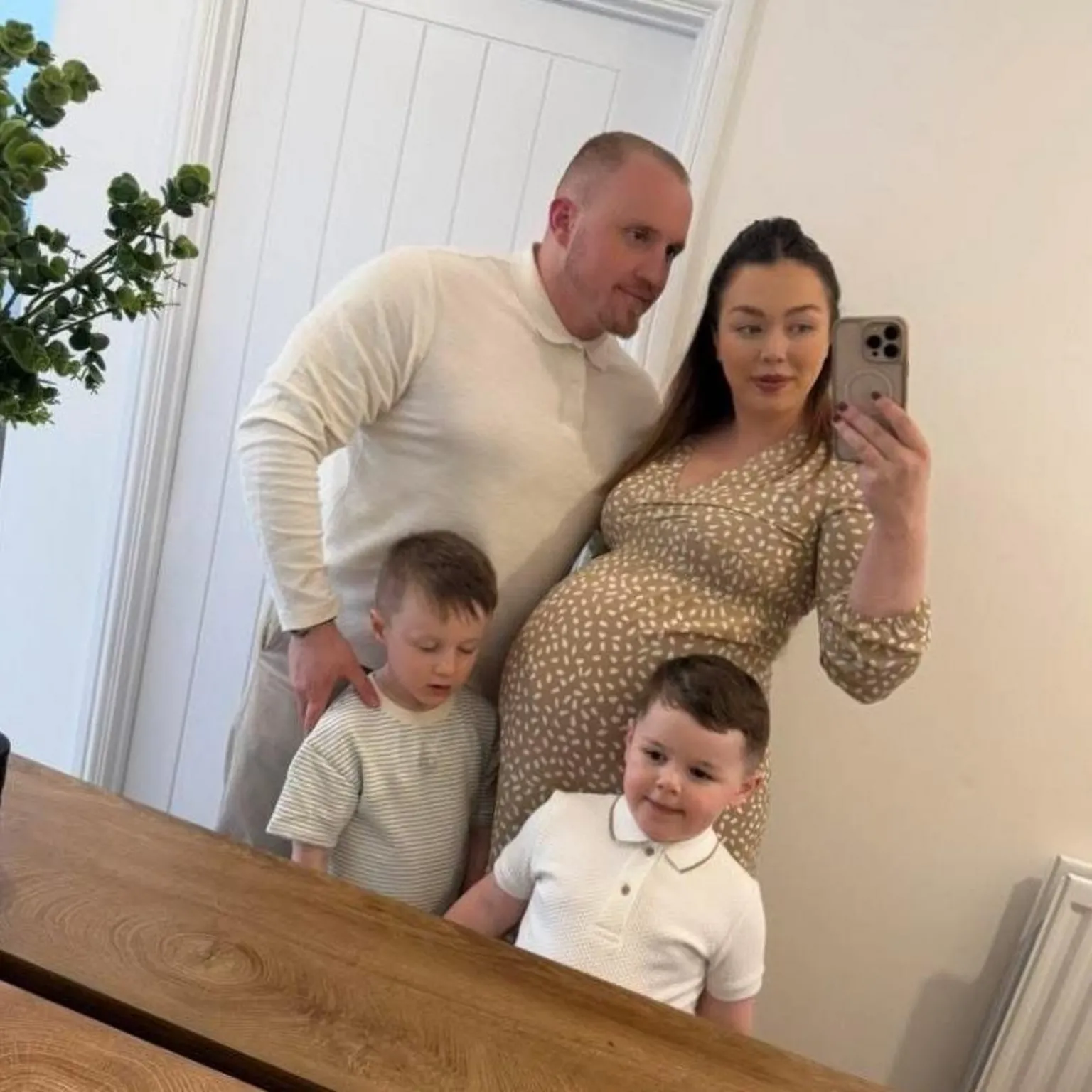 Keenan Acton A selfie picture of a man, a woman and two young boys in front of a mirror. The wall and door behind them are white, there is a brown wooden table in front of them and there is a plant in the corner. The man and two boys all have short fair hair and are wearing white tops. The woman has long brown hair, is wearing a brown and white patterned dress, has a baby bump and is holding a phone on one hand with her other arm around the man.