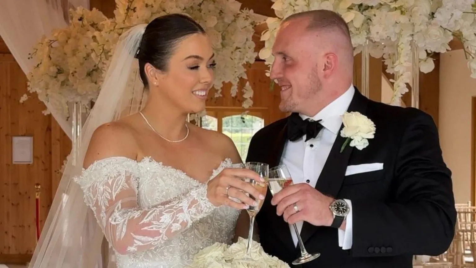 @thesocialwedit Keenan and Olivia on their wedding day. Olivia's dark hair is tied up off her face and she is wearing a white off-the-shoulder lace wedding dress and a long sheer veil. Keenan is wearing a black tux, black bow tie and a white shirt with a white rose button hole. They are toasting with champagne flutes at smiling at one-another.