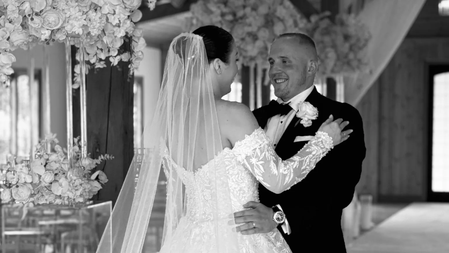 @thesocialwedit A black and white photo of Keenan and Olivia on their wedding day. Olivia's dark hair is tied up off her face and she is wearing a white off-the-shoulder lace wedding dress and a long sheer veil. Keenan is wearing a black tux, black bow tie and a white shirt with a white rose button hole. They are facing one another, Keenan has his hands on Olivia's hips and they are smiling.