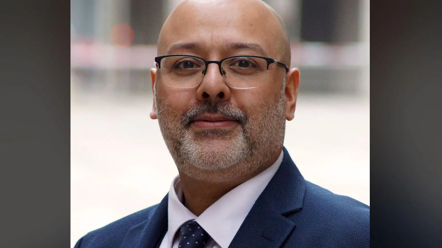 City of London Police Det Ch Insp Nik Jethwa is bald and has glasses, the head-and-shoulders shot shows him wearing a dark blue suit over a white shirt and a navy tie with white spots.