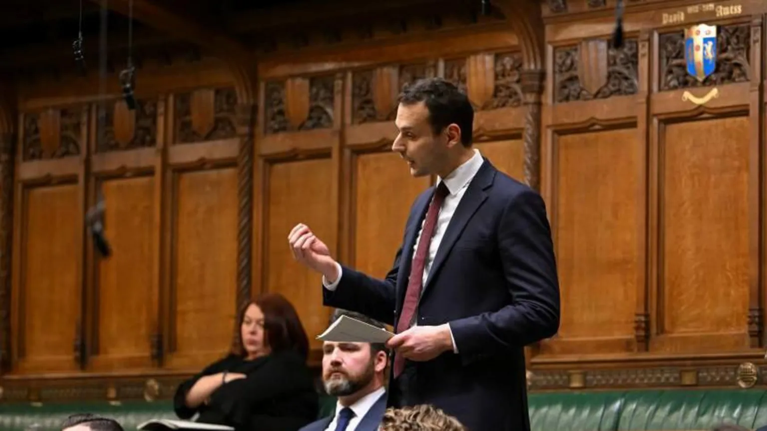 Labour MP Josh Simons speaks in the House of Commons. He is wearing a dark blue suit, burgundy tie and white shirt. He holds a piece of paper in his hand and gesticulates with his hand as he speaks. Other MPs sit on the green benches around him. 