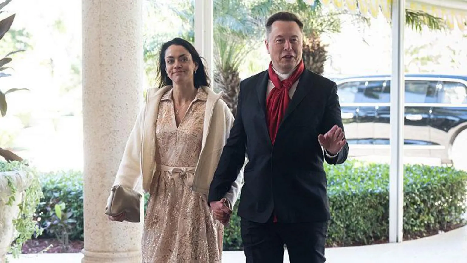  Shivon Zilis holding hands with Elon Musk. Zilis is wearing a pale dress and jacket, and Musk is wearing a suit and a red scarf. Both are smiling.