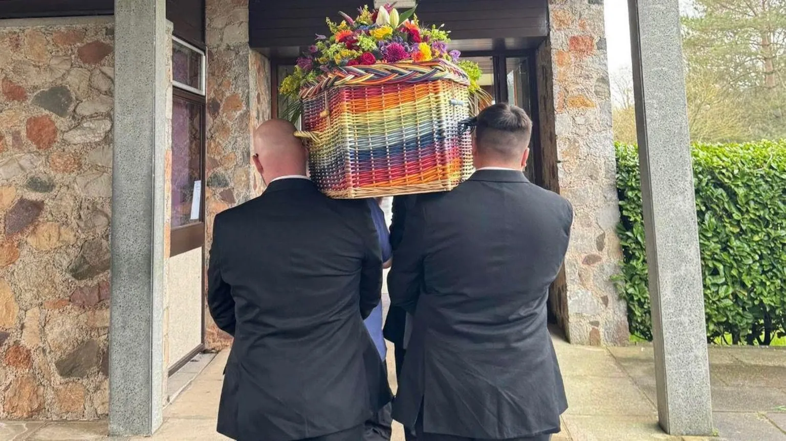 Simon Helliar-Moore A group of people carrying a multicoloured wicker coffin on their shoulders. They are walking as if in a procession and have their backs to the camera. They are wearing suits.