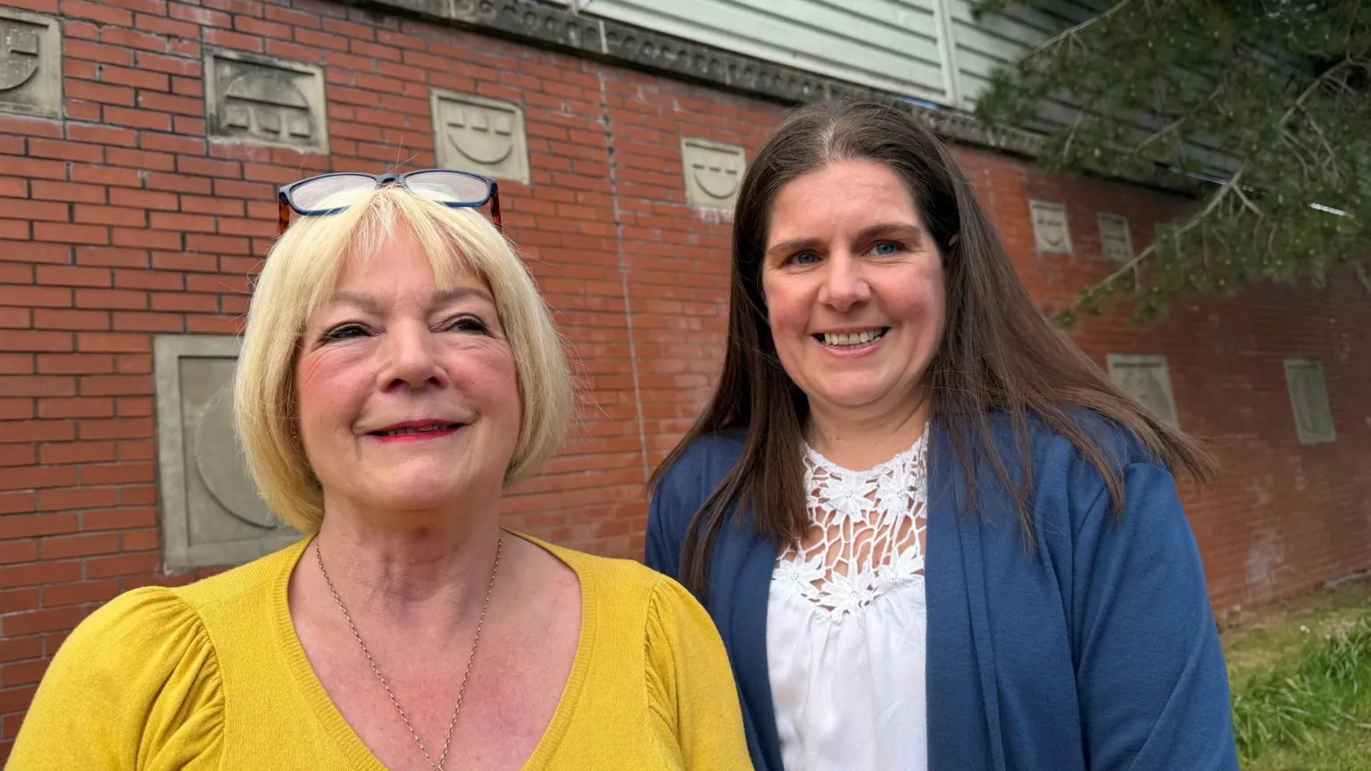 Two women stand side by side in front of a red brick wall. The one of the left has blonde hair, has reading glasses pushed onto her head and is wearing a mustard top. On the right the woman wears a blue cardigan over a white top which separates into a floral pattern around the neck.