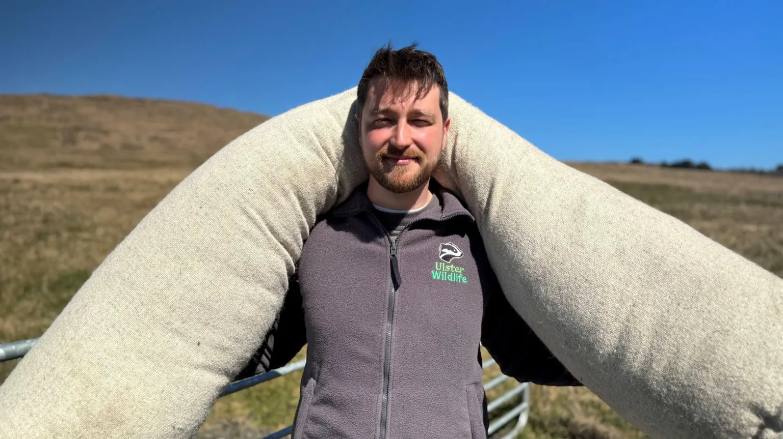 James Devenney is a young man with brown hair, brushed over his forehead. He has a short brown bears. He is wearing a grey fleece zip up top with the Ulster WIldlife badger logo on it. He is carrying one of the wool logs over both his shoulders. Behind him is blue sky and peatland. 