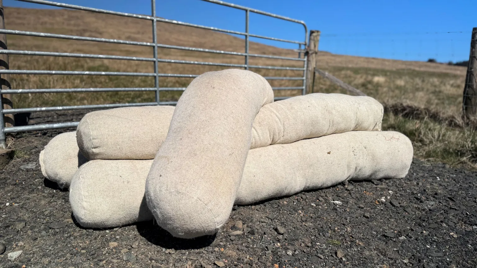 A pile of wool logs sit on the ground in front of a gate leading to a field. They are long and cylindrical and have an outer covering made of a linen-coloured woollen textile. 