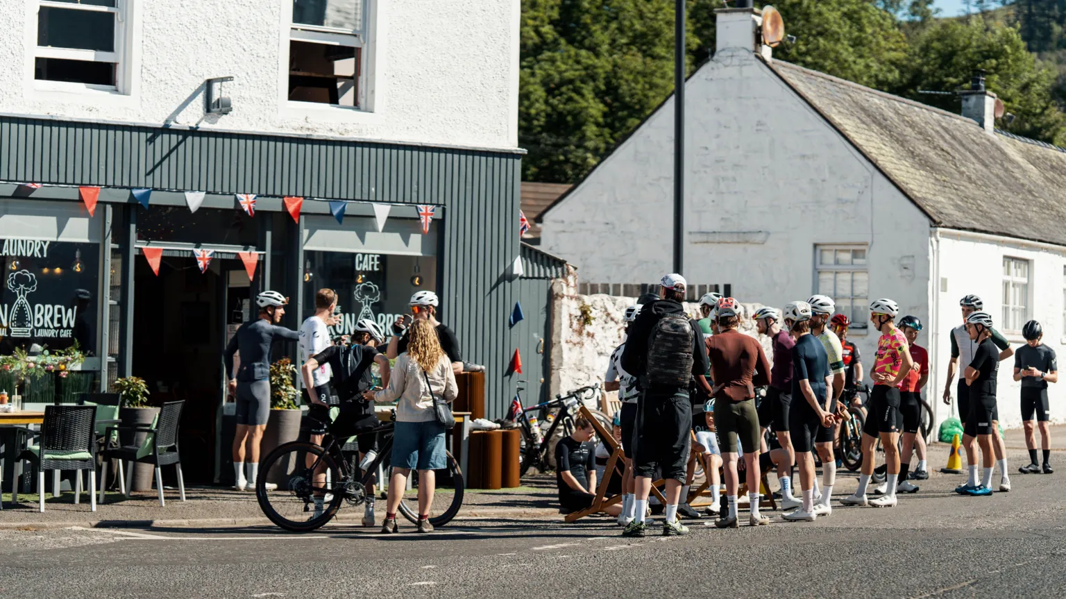 Red On Sports A group of cyclists gathered outside a cafe, most of them are standing without their bikes