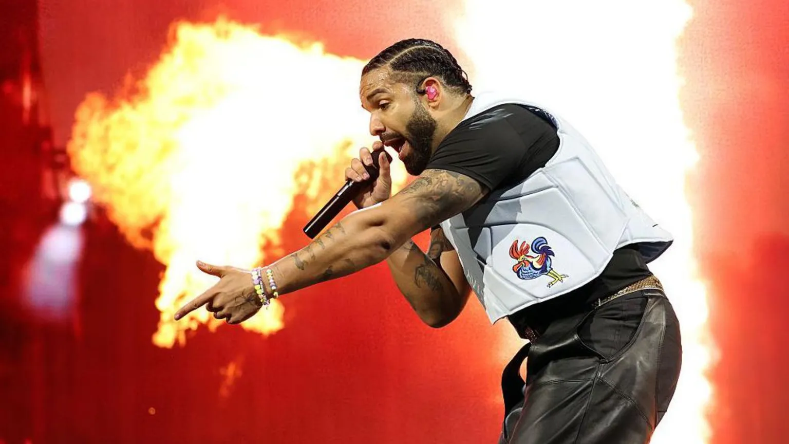  Drake performs on stage as a plume of flame erupts behind him, framing the star as he raps into a microphone and points into the crowd with his free hand. Wearing a white vest with a cartoon image of a bird printed on it, he looks intense.
