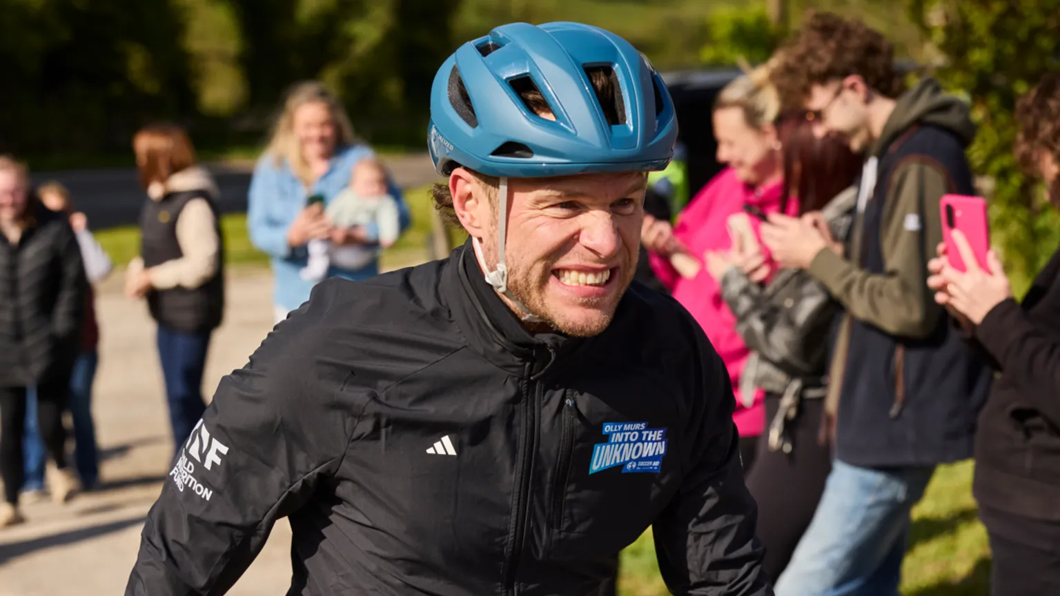 Unicef Olly Murs with a clenched face and stubble, wearing a cornflower blue helmet. His top is visible and bears the logo of the challenge he is doing, called Into The Unknown Challenge. The brown curls of his short hair are just visible by his left ear. Blurred in the background are the spectators, holding their phones and smiling as Murs pedalled through the Hope Valley
