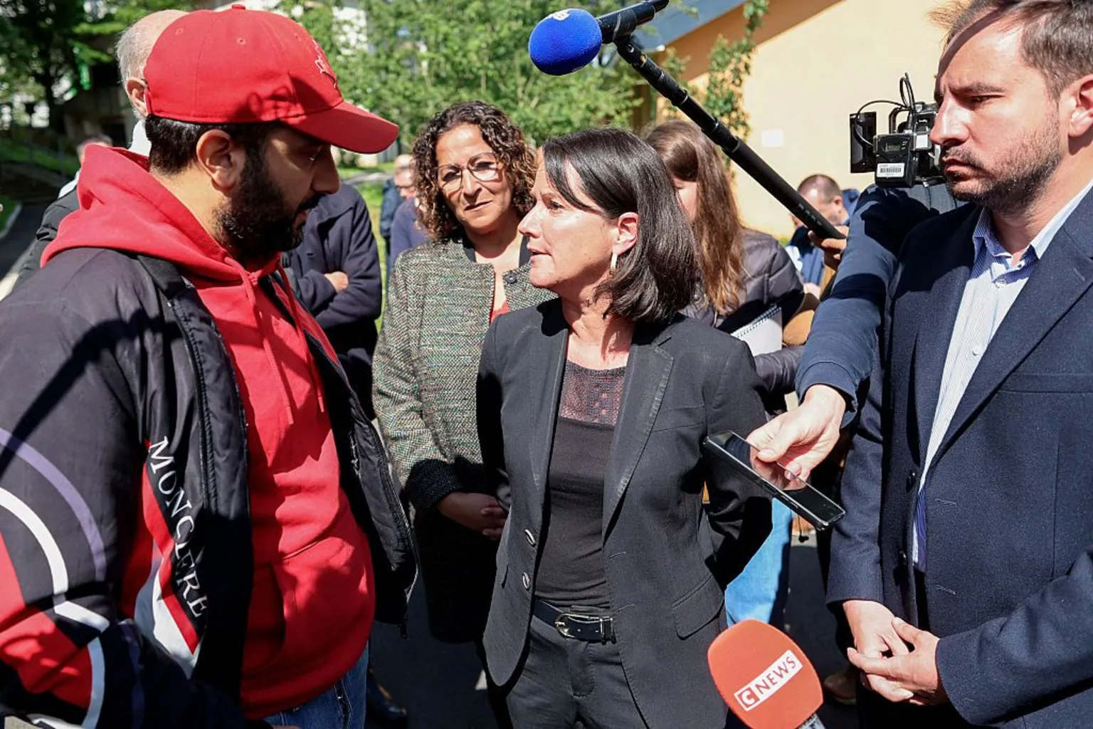  Local mayor Johanna Rolland (C), wearing a black jacket, top and trousers, speaks to a man wearing a red cap and hoodie and a dark jacket. A microphone is held above her head and several other journalists are seen recording her remarks with phones and microphones.