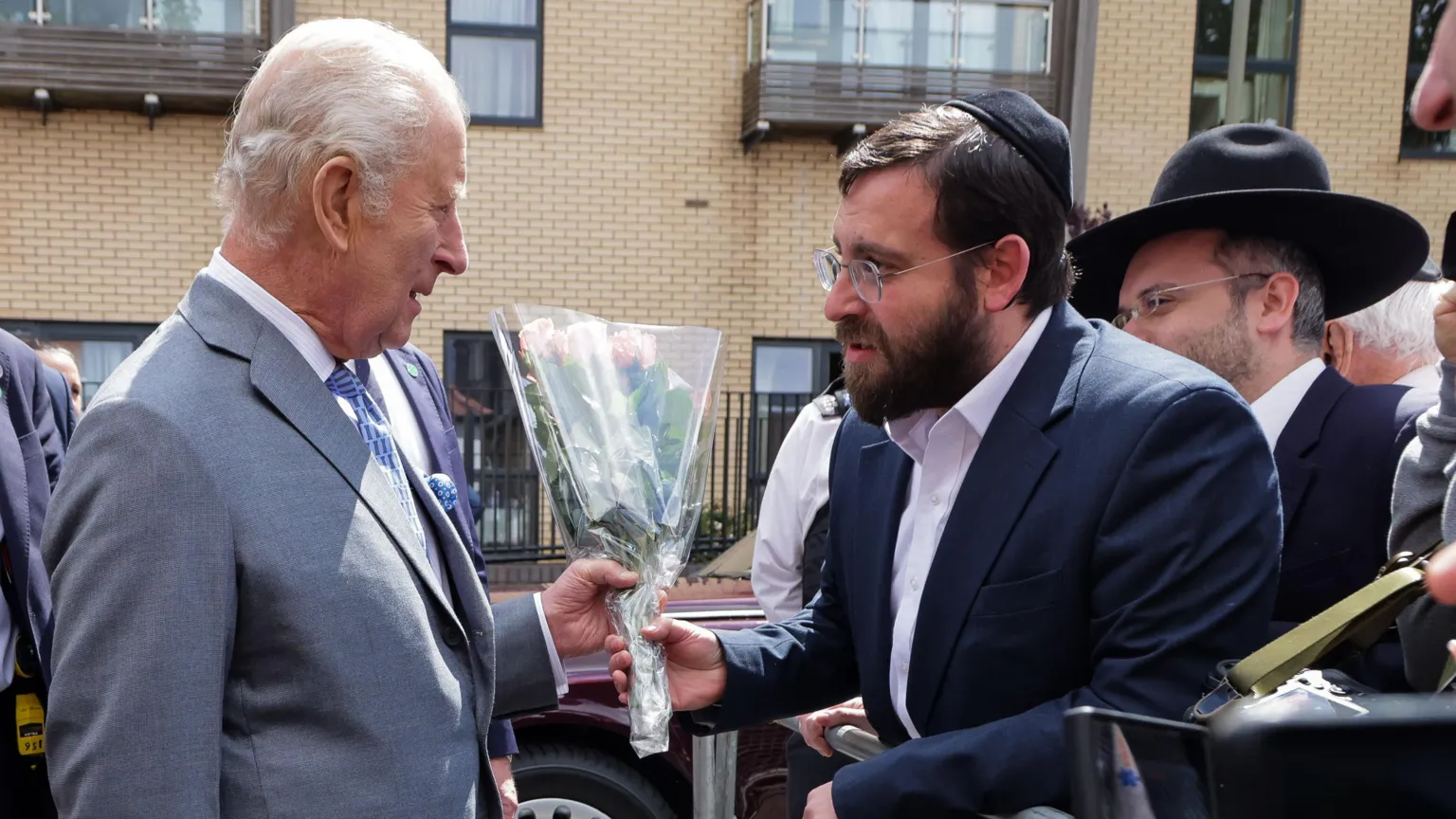  King Charles III in a grey suit is presented with flowers by a Jewish member of the community of Golders Green who is standing behind a barrier with a crowd of other Jewish people.