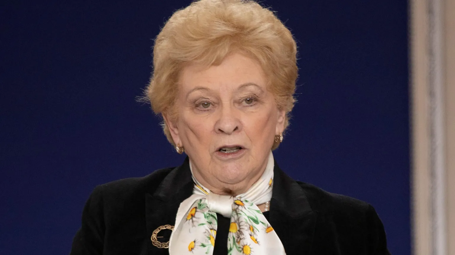  Nuala O'Loan in black jacket with blonde hair and white scarf with floral pattern.