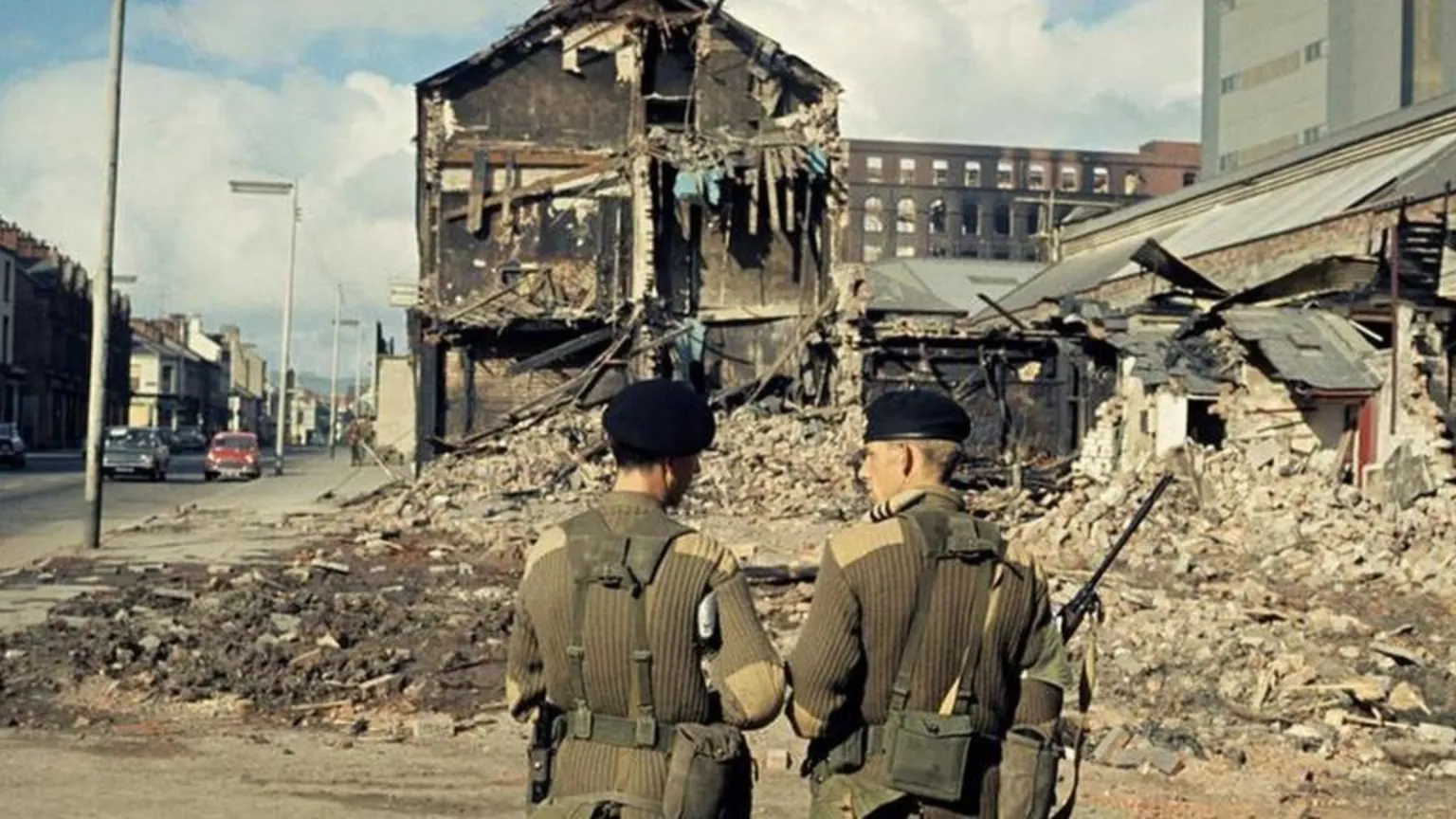 An archive photo of two British soldiers in green uniforms and black berets with their backs to camera, a bombed building is visible in front of them with debris all across the road.