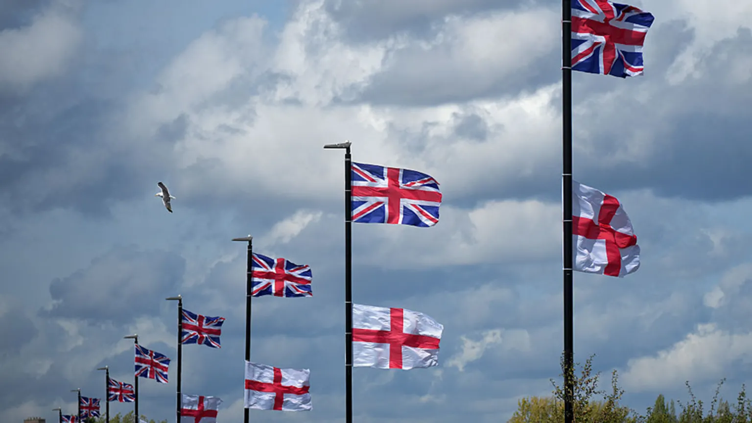  Lamp-posts with union jacks flying from the top, with England flags slightly below them. A cloudy sky is in the background