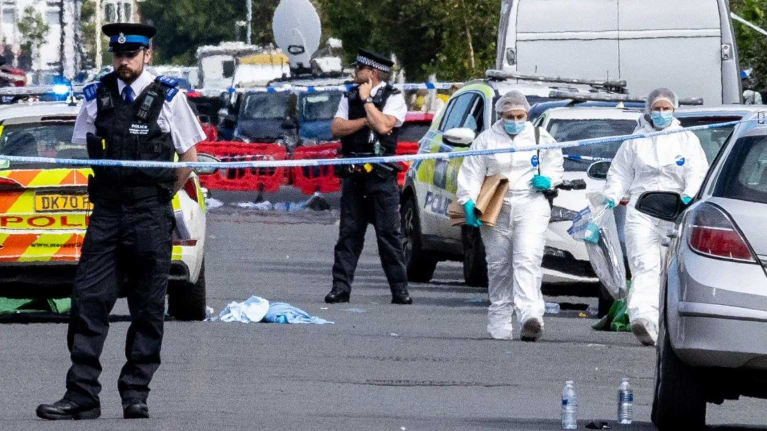  Two forensics are walking behind police tape between parked cars. Two police officers are standing behind the cordon. 