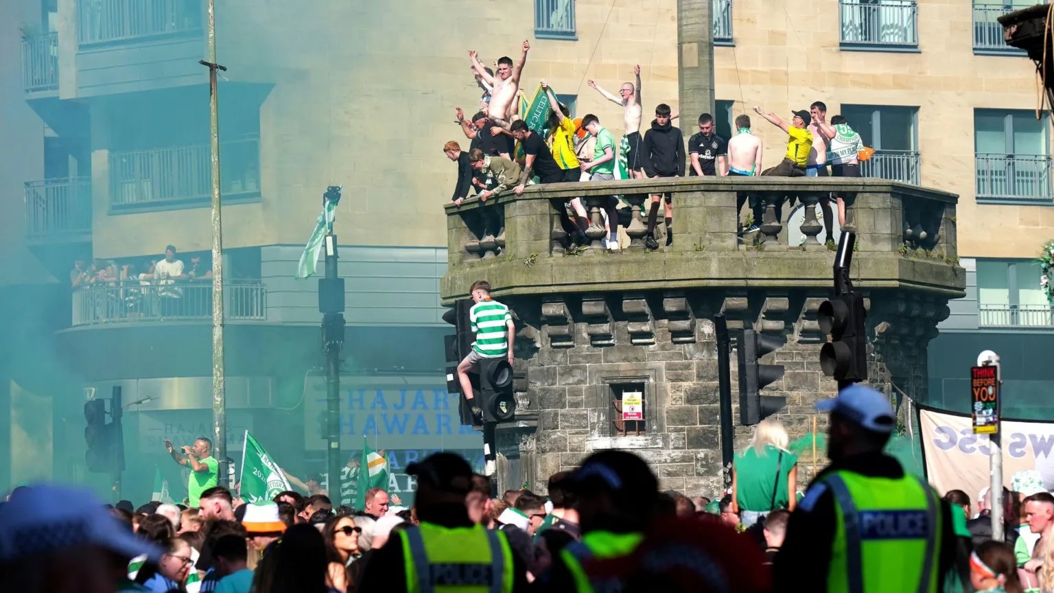  Celtic supporters, some wearing green and yellow football tops, gathered in celebration on top of the city's historic Mercat Cross. The crowd includes two bare-chested men whose arms are rasied in triumph. Another supporter - wearing green and white hoops - is sitting on top of a set of traffic lights as crowds mill in the street below. Uniformed police officers look on.