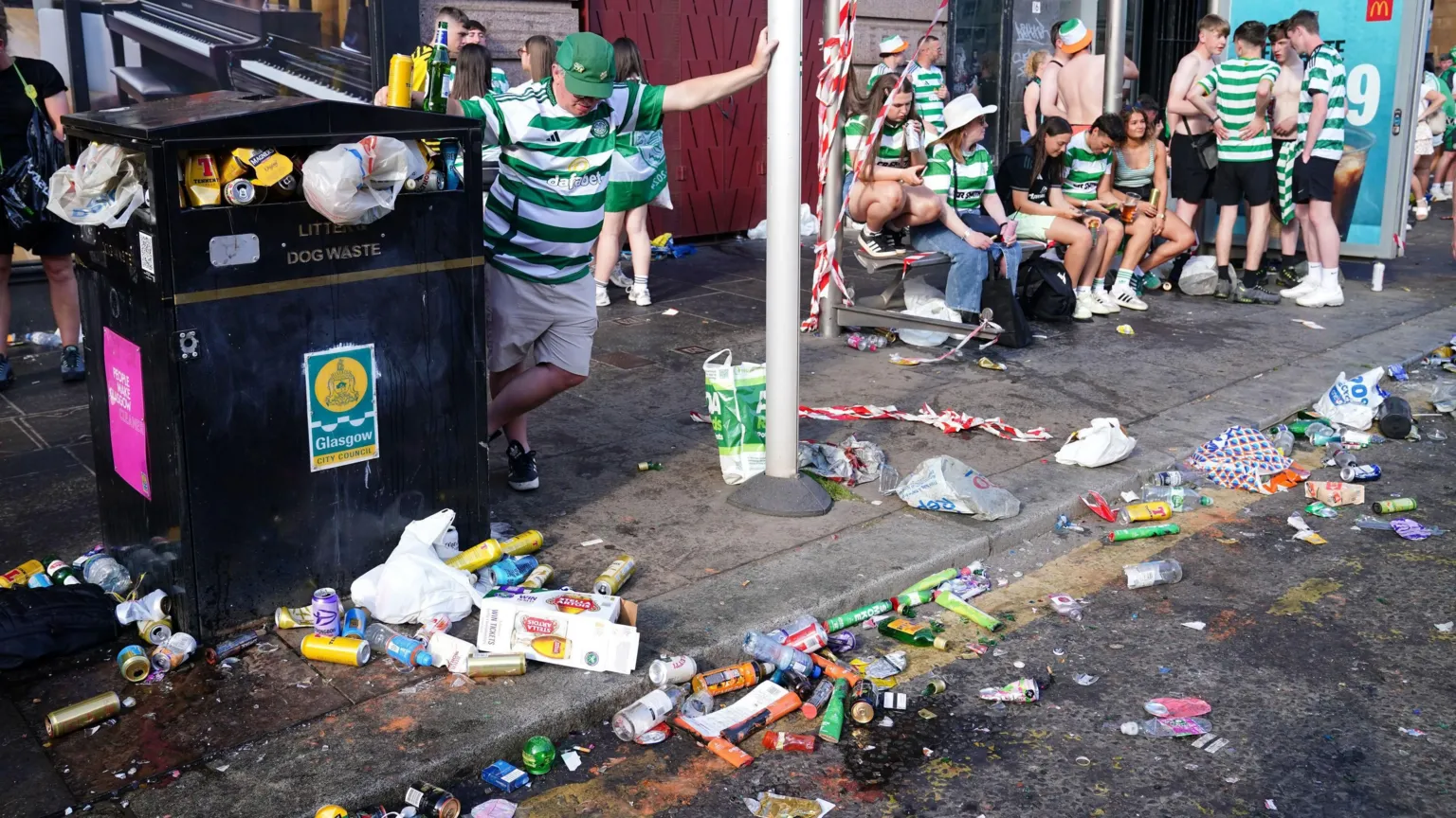  A Celtic fan wearing a green cap and green and white hoops, uses a bus stop to support himself. A crowd of young people - also wearing Celtic colours - sit on an adjoining bench. A nearby bin is overflowing and the street is strewn with rubbish, including empty beer bottles and cans.