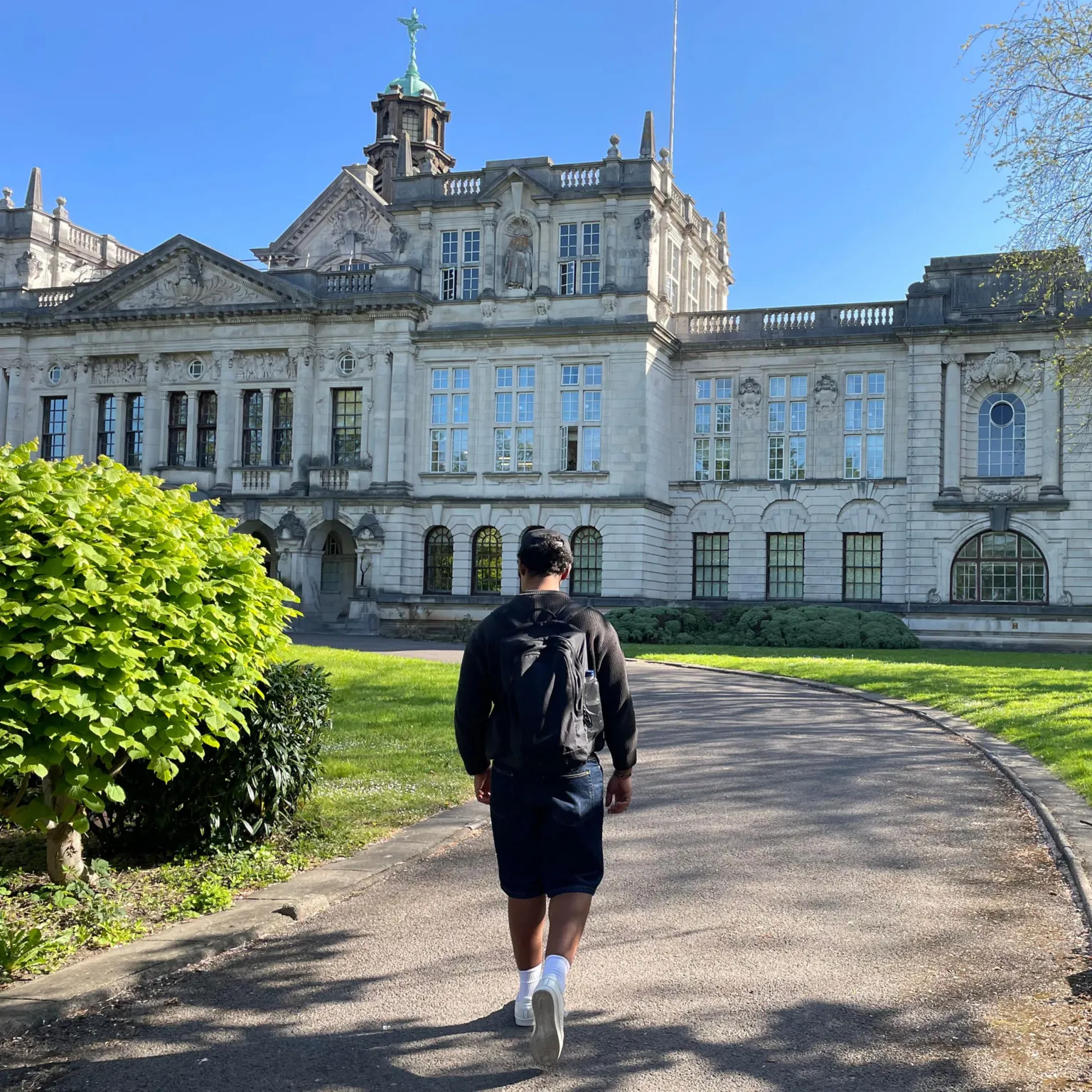 Jordan walks away from the camera along a curved path toward Cardiff University main building. A large, ornate stone building with arched windows, columns, and decorative stonework. A stone pillar or wall edge appears on the right side of the frame. The lawn is neatly trimmed and trees cast long shadows in bright sunlight. Jordan is wearing denim shorts, white trainers, a black rucksack and a black cap. He has black curly hair. 