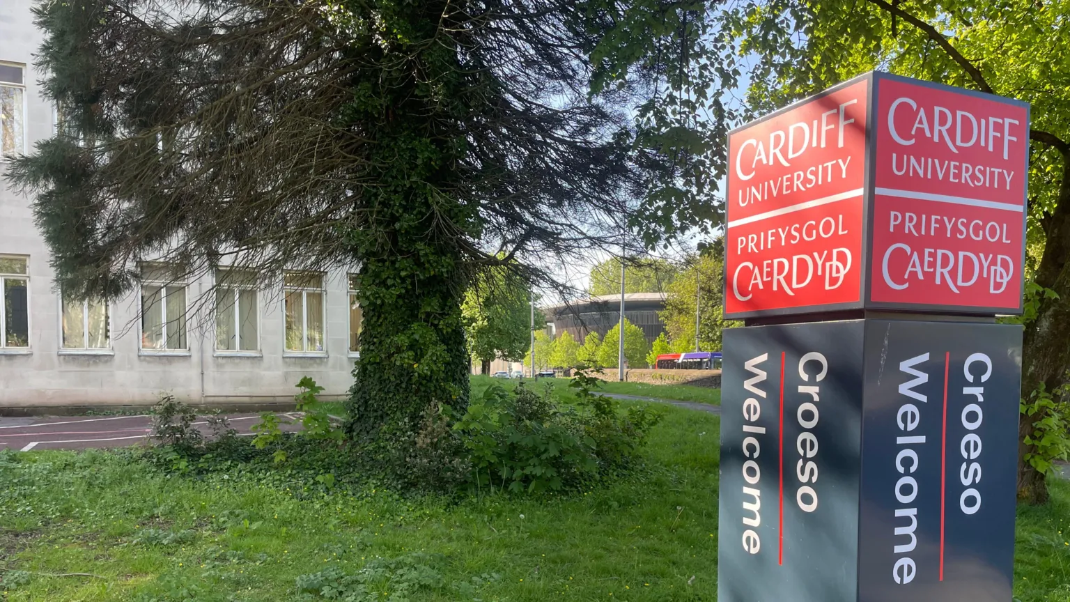 A tall rectangular sign stands on grass beside trees and a path. The sign reads “Cardiff University / Prifysgol Caerdydd” in white text on a red background, with “Croeso / Welcome” printed vertically below on a dark panel. Behind the sign is a light‑coloured stone building partly obscured by trees and foliage.