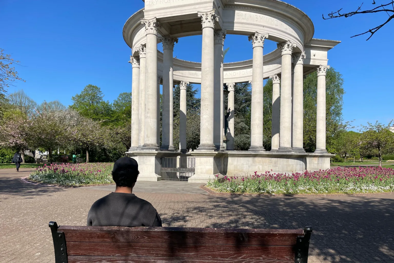 In the foreground, a wooden park bench fills the lower frame, with Jordan seated facing away from the camera. Beyond the bench is a wide paved path leading to a large circular stone pavilion supported by evenly spaced columns. The pavilion is surrounded by neatly planted pink flowers, with tall green trees behind it. 