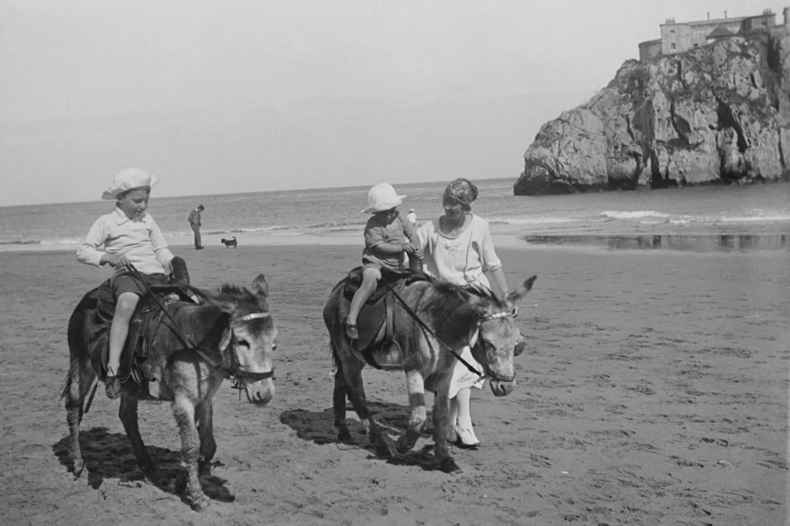  A woman leads walks alongside two donkeys carrying small children on Tenby's South Beach in 1924 in a black and white photo. Shewears a long white dress. The children are wearing shorts, shirt and sunhat and a dress and a sunhat. A man and a dog are visible in the background at the water's edge.