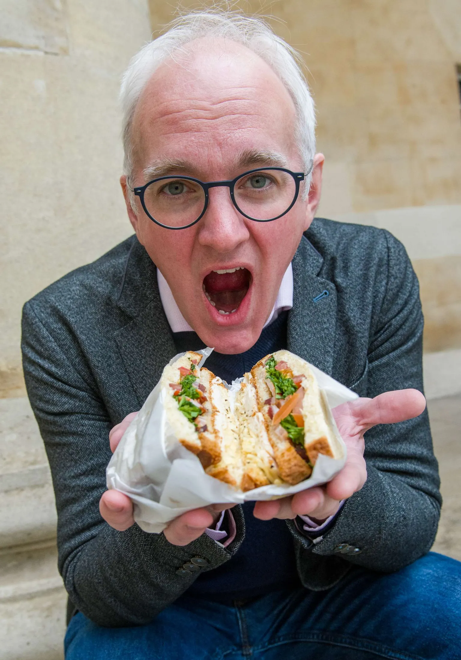 Emma Lynch/BBC The image shows a close-up, centered view of a person seated against a light stone background, holding a sandwich wrapped in white paper with both hands. The sandwich is cut in half, revealing multiple layers including sliced meat, melted cheese, leafy greens, and pieces of tomato inside toasted bread. The person is wearing round, dark-framed glasses along with a textured jacket layered over a collared shirt and a dark sweater. The sandwich is positioned prominently in the foreground, with its filling clearly visible.