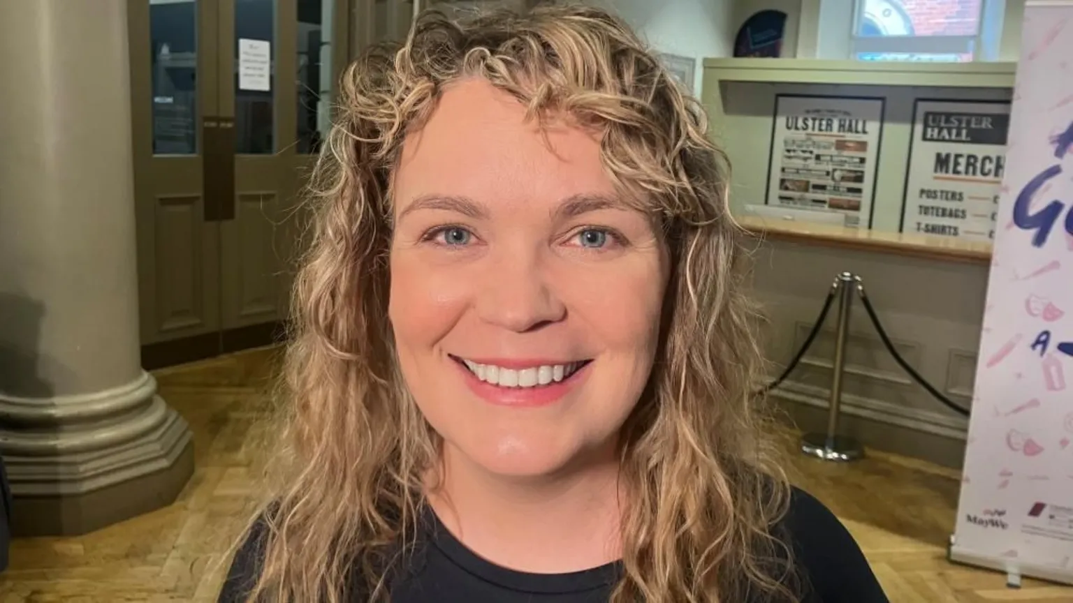 Has curly blonde hair and is smiling at the camera. She is wearing a black top.