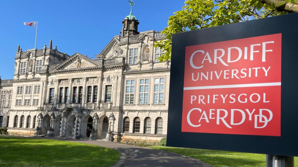 Mae arwydd coch yn nodi “Cardiff University / Prifysgol Caerdydd”. Y tu ôl iddo, mae llwybr llydan yn troi tuag at yr adeilad prifysgol.