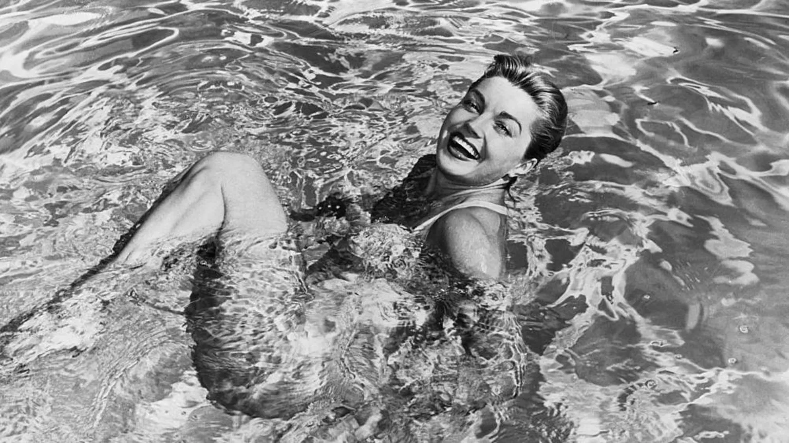 Esther Williams in a swimming pool. She is on her back with her knees bent and head turned towards the camera, smiling. 