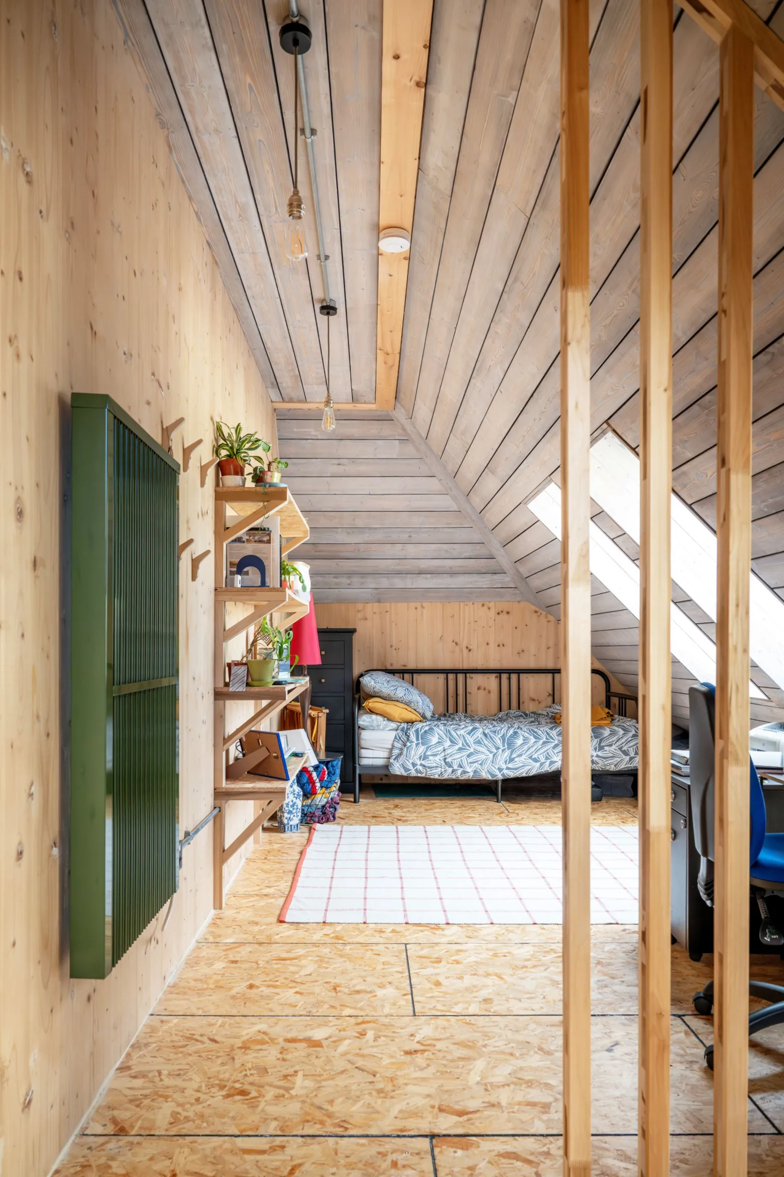 IWC Media More timber in a view through an opening into a bedroom, lined in wood, with a wooden floor and wood shelving. Bright skylights on the sloped right hand side flood the room with light.